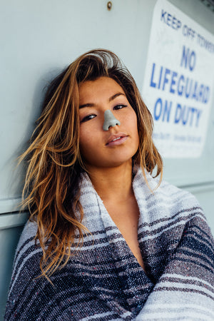 Young lady wearing Nöz blue colored reef safe eco friendly sunscreen on nose while leaning on lifeguard tower at the beach. 